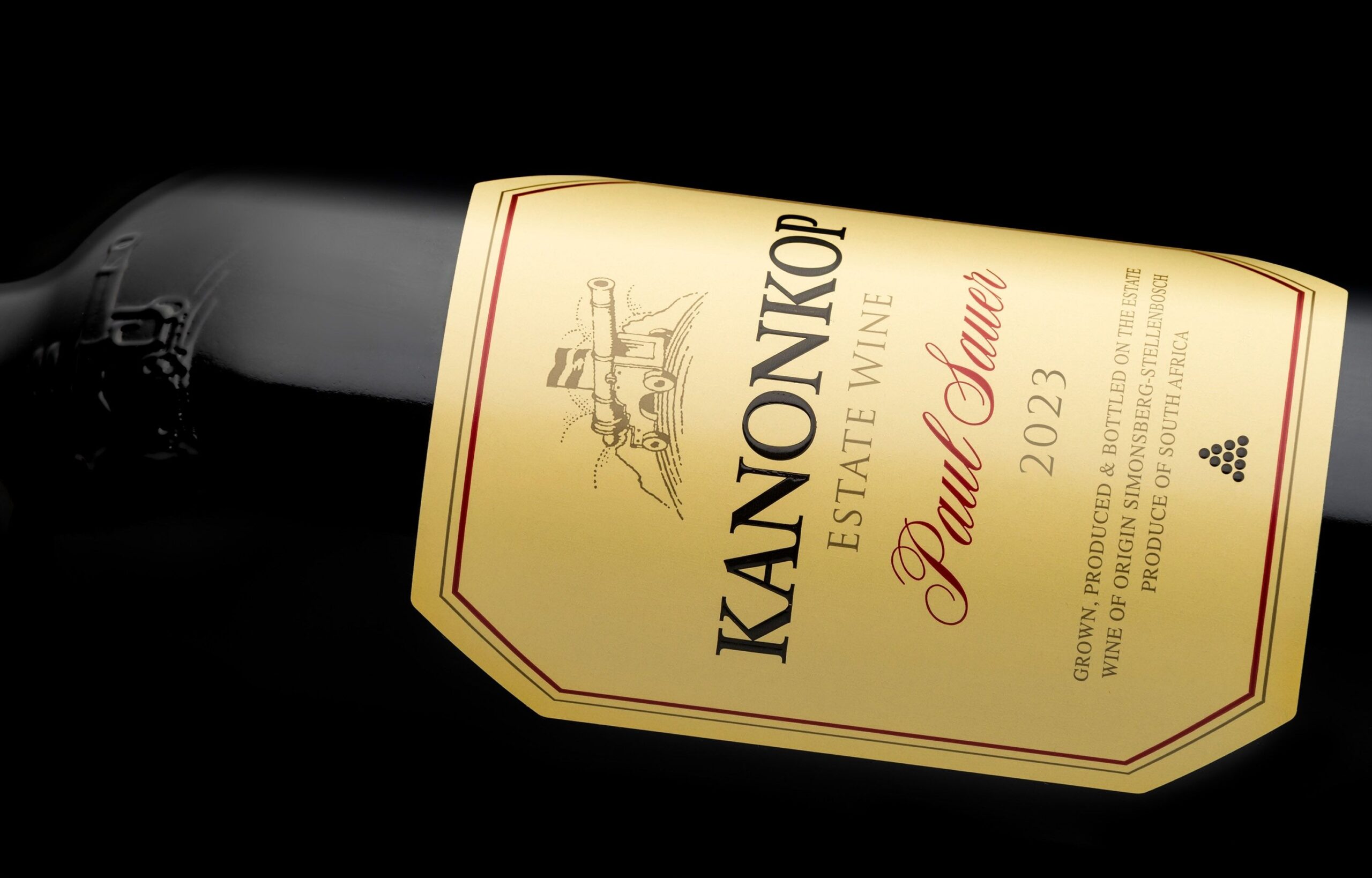 Styled bottle shot of the new Kanonkop Paul Sauer 2023 wine, leaning to the left with its label clearly visible, facing upwards. A black background places focus on the words written on the label “ Kanonkop Estate Wine, Paul Sauer, 2023”, and in smaller text “Origin, produced, and bottled on the estate. Wine of Origin Simonsberg-Stellenbosch. Product of South Africa”