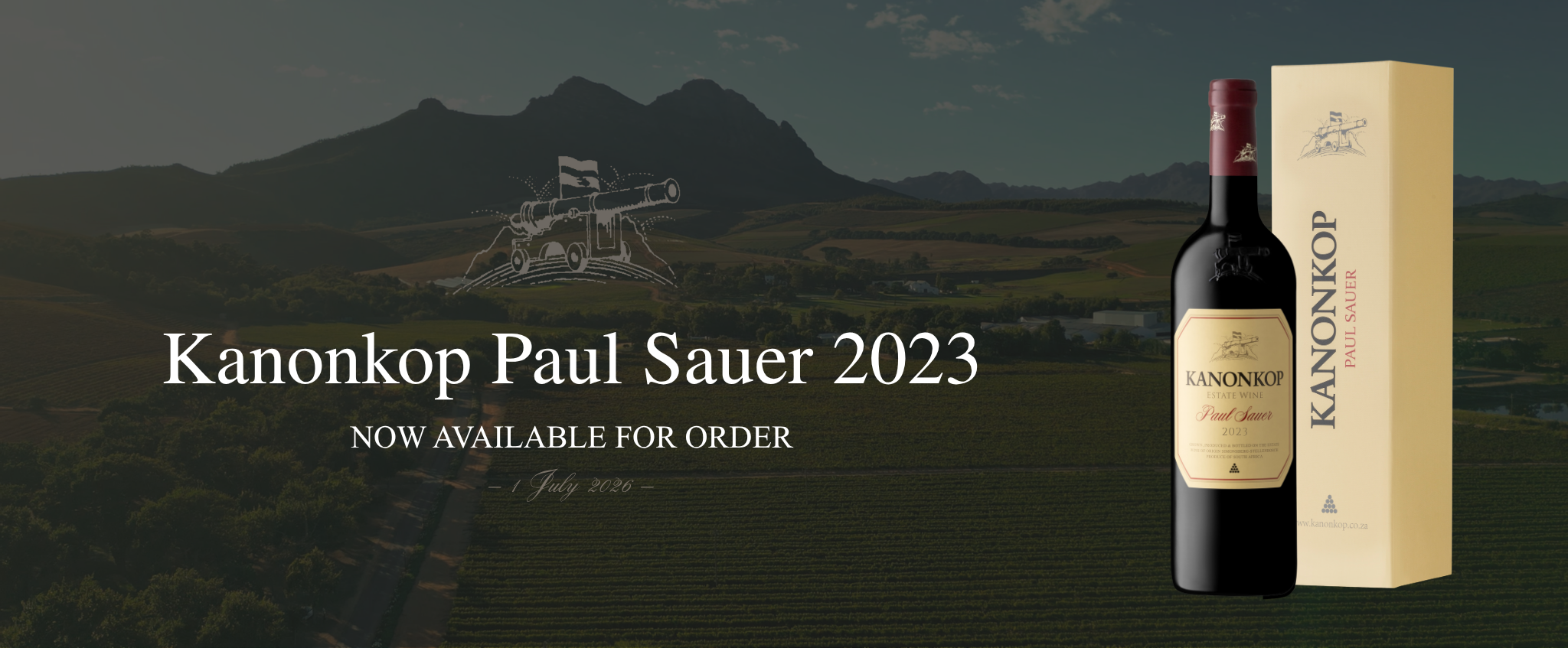 A promotional banner for the release of Kanonkop Paul Sauer 2023 wine. The background features a darkened panoramic view of the Kanonkop Wine Estate vineyards at dusk, with rolling rows of vines leading toward a dramatic Simonsberg mountain. Centred near the top is a subtle illustration of the Kanonkop cannon logo. Large white text reads "Kanonkop Paul Sauer 2023," with smaller capitalized text beneath stating "Now available for order." Below this, in elegant script, appears the date "1 July 2026." On the right side of the banner, a bottle of Kanonkop Paul Sauer 2023 stands beside its vintage-cream presentation box, both prominently displayed with the estate branding clearly visible. 